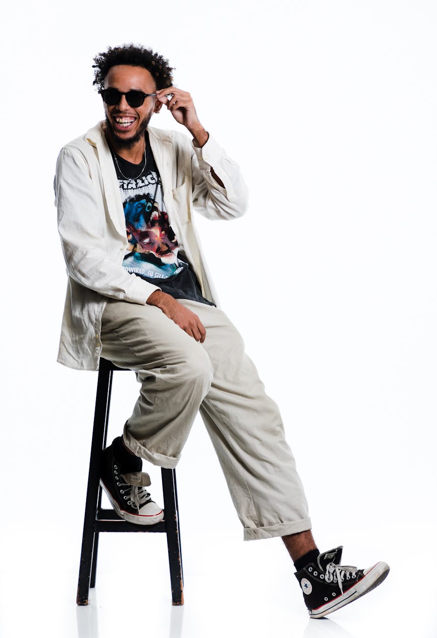 Casual portrait of a stylish young man wearing sunglasses and casual wear, seated on a stool.