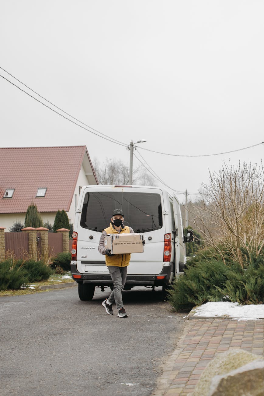 A delivery person wearing a mask carries a box from a van in a residential area.