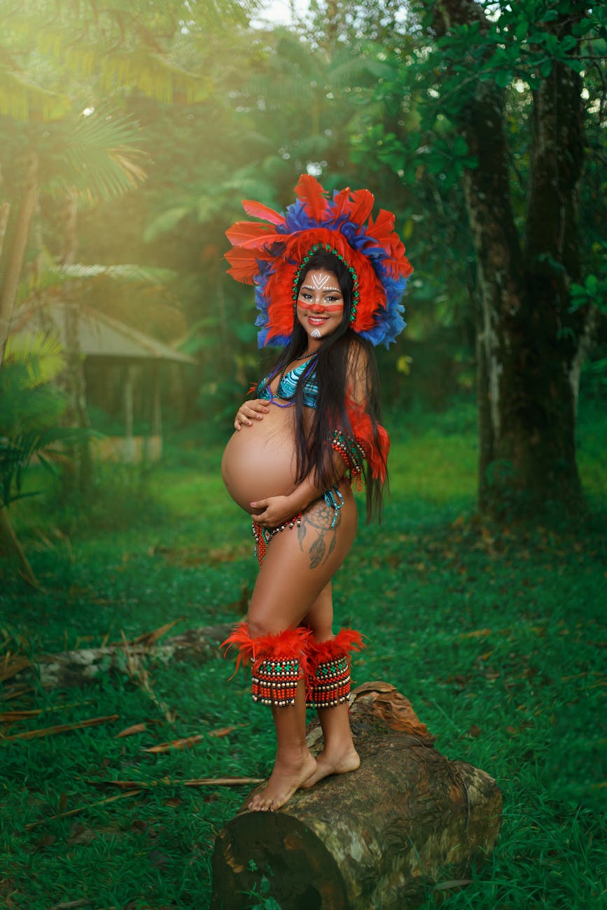 A smiling pregnant woman in traditional Brazilian attire, standing outdoors with tribal paint and headdress.